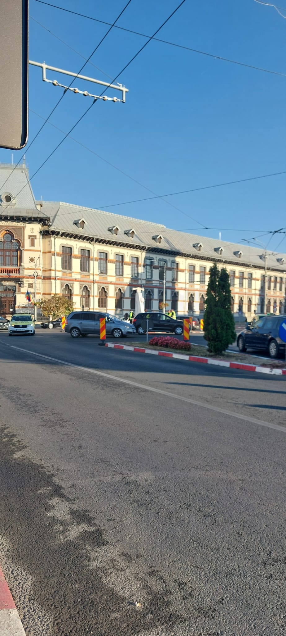 Historic city street with an ornate European-style building, multiple cars, and orange construction barriers under a clear blue sky.