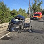 Wrecked dark blue sedan embedded against a highway barrier with debris on the road; a red emergency vehicle is approaching in the background.