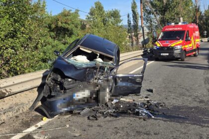 Wrecked dark blue sedan embedded against a highway barrier with debris on the road; a red emergency vehicle is approaching in the background.