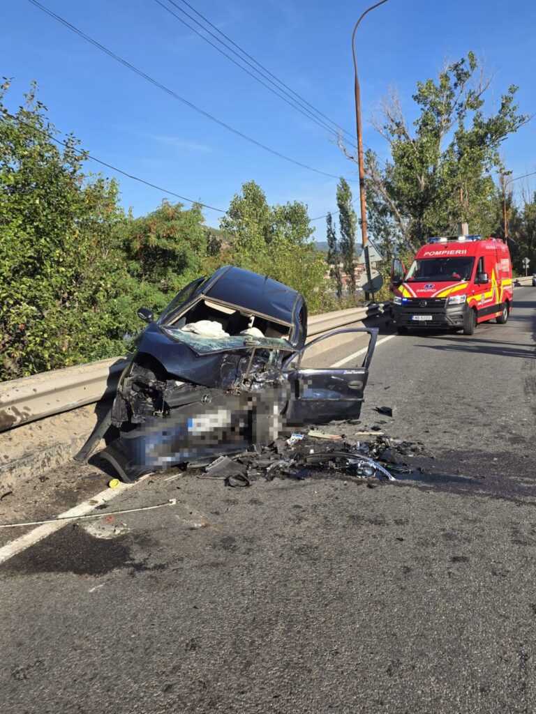 Wrecked dark blue sedan embedded against a highway barrier with debris on the road; a red emergency vehicle is approaching in the background.