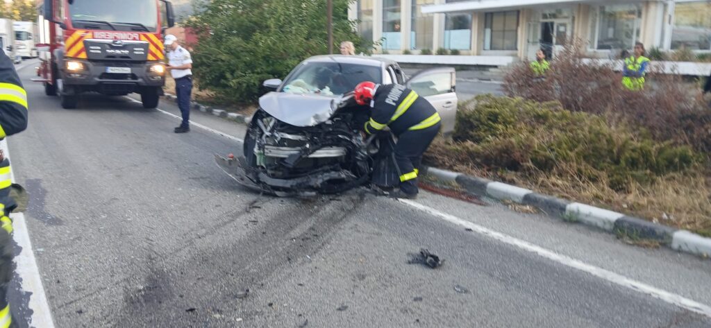 Car crash on a street with front-end damage; firefighter kneels by the wreck while emergency responders observe nearby.