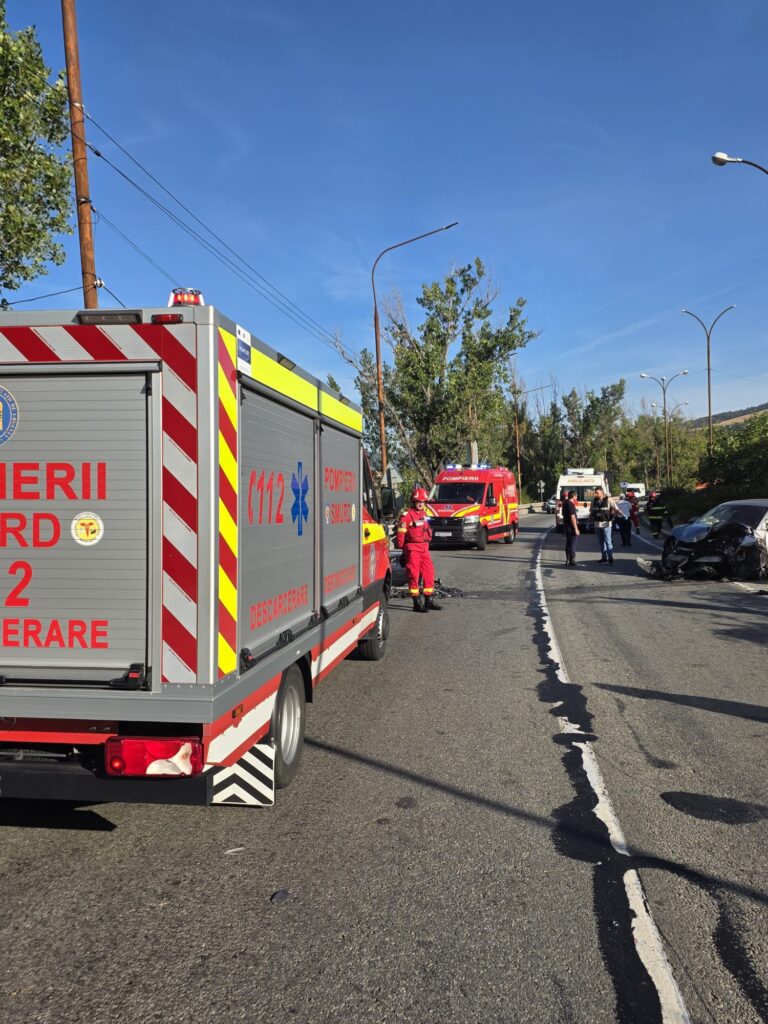 Fire trucks and emergency responders on a sunny road; a damaged black car visible on the right side of the scene.