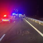 Nighttime highway crash scene with police car, ambulance, and firefighters tending to an incident in the distance with flashing lights.