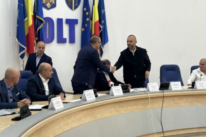 Two men in suits shake hands across a curved conference table during a formal meeting, with flags and emblems in the background.