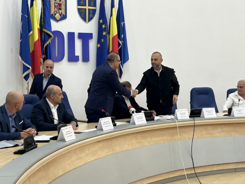 Two men in suits shake hands across a curved conference table during a formal meeting, with flags and emblems in the background.