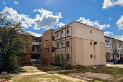 Construction scaffolding against a pale apartment building under a bright blue sky with scattered clouds and trees nearby, with debris on the ground.