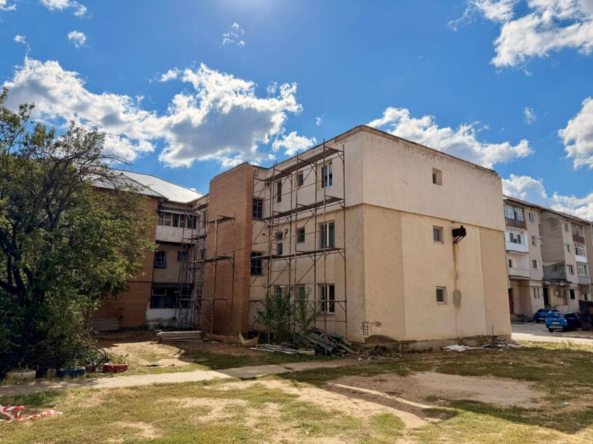 Construction scaffolding against a pale apartment building under a bright blue sky with scattered clouds and trees nearby, with debris on the ground.