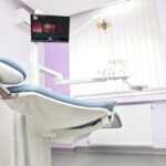 Dental clinic treatment chair in a pale purple-walled room with attached overhead light and tools in the background.