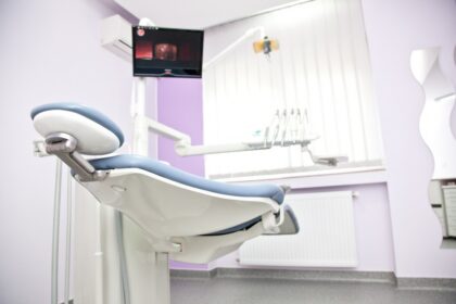 Dental clinic treatment chair in a pale purple-walled room with attached overhead light and tools in the background.