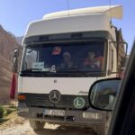 White Mercedes truck on a dusty mountain road; two people visible in cab; Romanian plate OT 25 GYN; green 'L' learner sign on bumper.