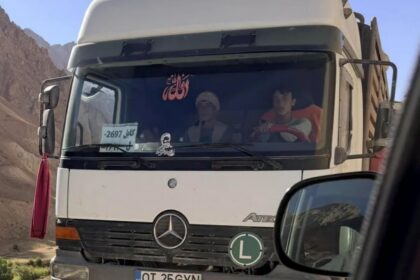 White Mercedes truck on a dusty mountain road; two people visible in cab; Romanian plate OT 25 GYN; green 'L' learner sign on bumper.