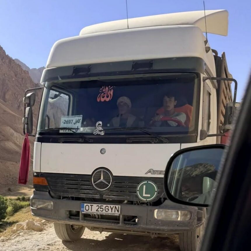 White Mercedes truck on a dusty mountain road; two people visible in cab; Romanian plate OT 25 GYN; green 'L' learner sign on bumper.