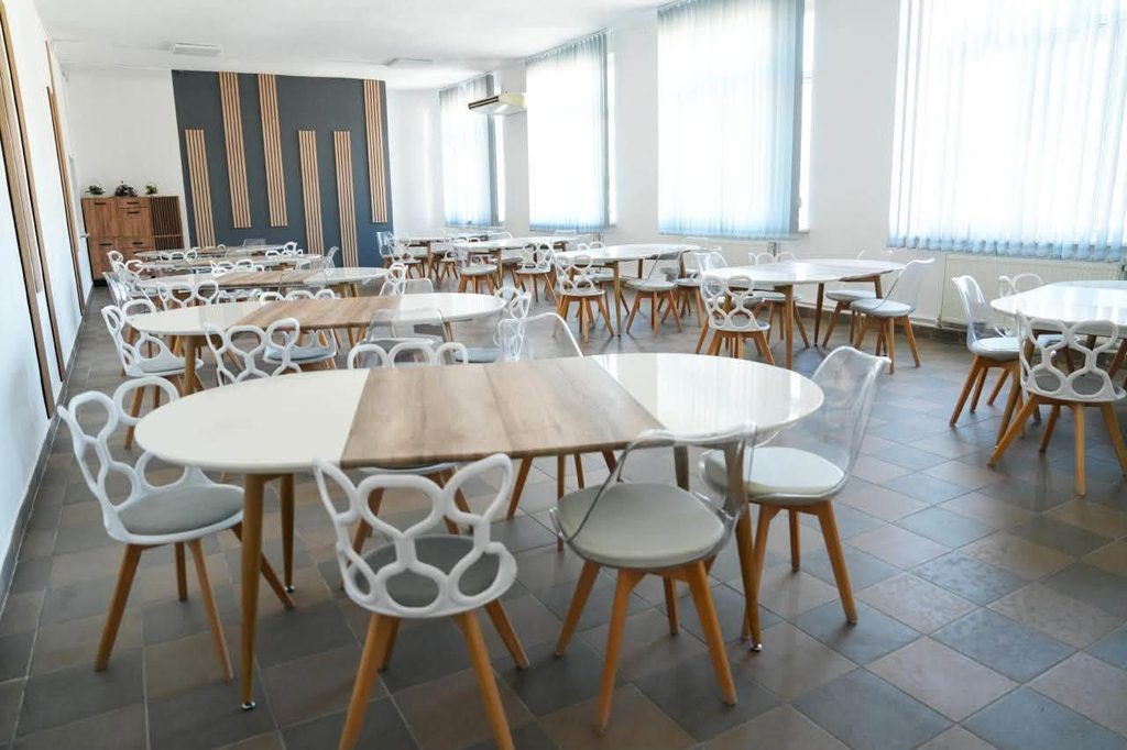 Bright cafeteria/dining area with round white tables and white molded chairs, wooden legs, and large windows with vertical blinds.