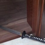 A Japanese katana with a black-wrapped handle lies on a tiled floor near a wooden door frame, blade extending under the door."