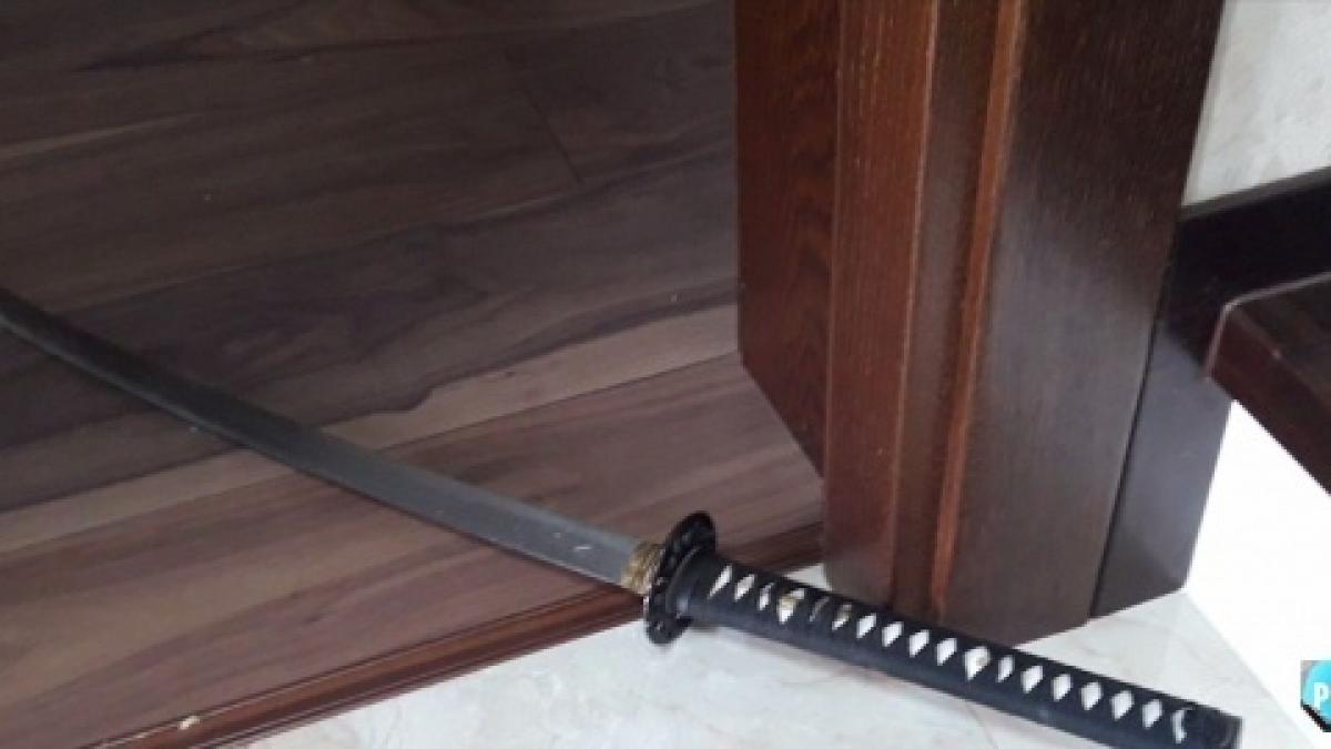 care-mai-este-starea-politistului-atacat-cu-sabia-498049 - Jurnalul Olteniei – Știri din Craiova și Oltenia A Japanese katana with a black-wrapped handle lies on a tiled floor near a wooden door frame, blade extending under the door."