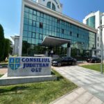 Modern government building with blue-tinted glass façade, entrance canopy, and a stone sign reading Consiliul Județean Olt in front.