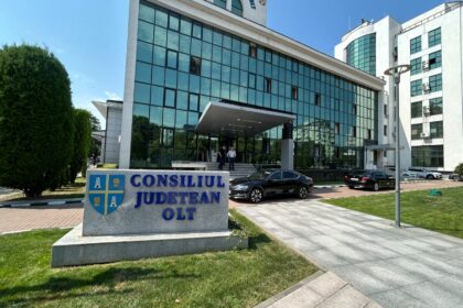 Modern government building with blue-tinted glass façade, entrance canopy, and a stone sign reading Consiliul Județean Olt in front.