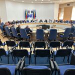Large round conference table with many attendees in blue chairs, flags and coats of arms on the wall behind.