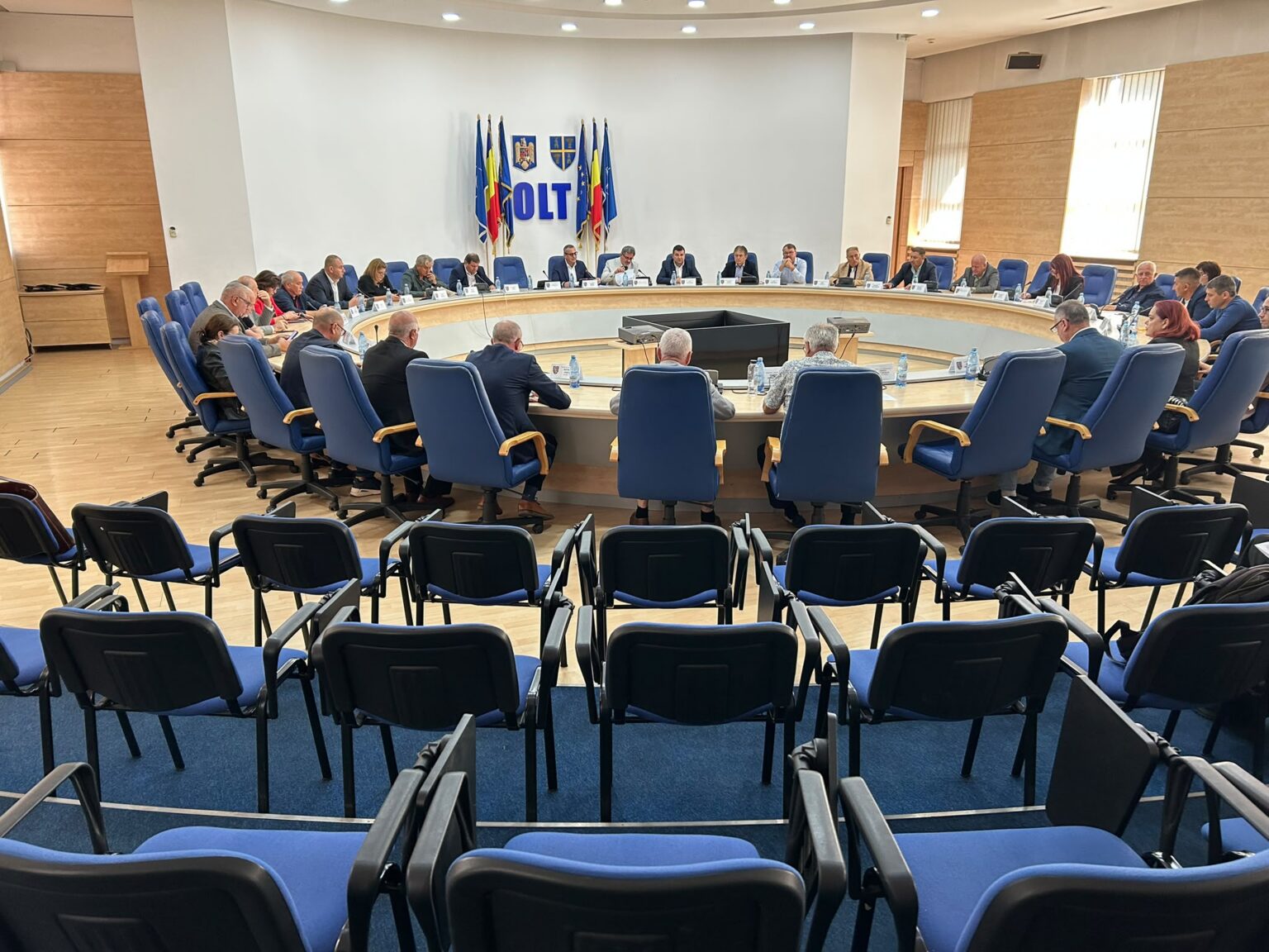 Large round conference table with many attendees in blue chairs, flags and coats of arms on the wall behind.