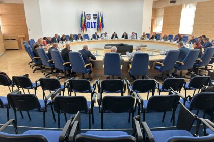Large round conference table with many attendees in blue chairs, flags and coats of arms on the wall behind.