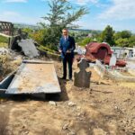 Man in a blue suit standing in a cemetery beside damaged gravestones and construction debris, with a village on the horizon.