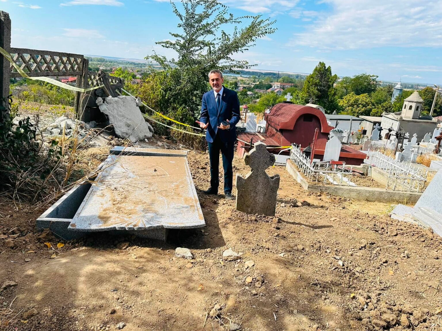 Man in a blue suit standing in a cemetery beside damaged gravestones and construction debris, with a village on the horizon.