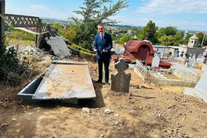Man in a blue suit standing in a cemetery beside damaged gravestones and construction debris, with a village on the horizon.