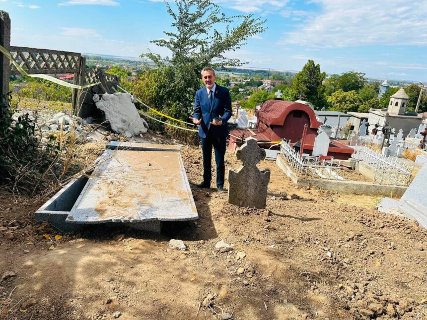 Man in a blue suit standing in a cemetery beside damaged gravestones and construction debris, with a village on the horizon.