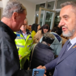 Two men shake hands in a crowded indoor event, with a police officer in a neon vest visible in the background.