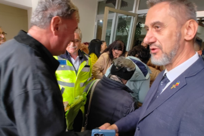 Two men shake hands in a crowded indoor event, with a police officer in a neon vest visible in the background.