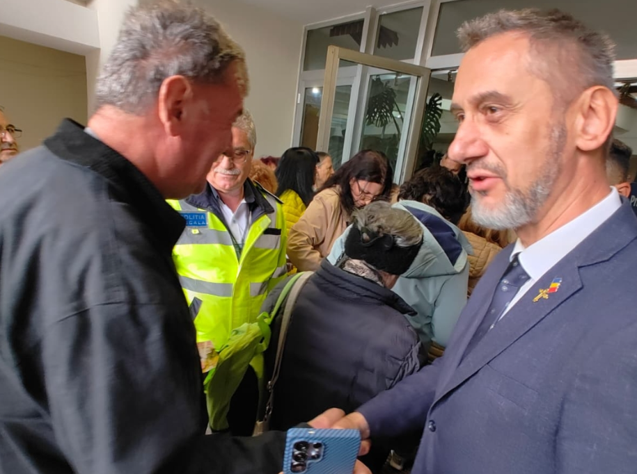 Two men shake hands in a crowded indoor event, with a police officer in a neon vest visible in the background.