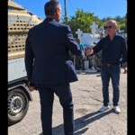 Two men shake hands beside a beige truck in a sunny cemetery with white crosses in the background, wearing smart and casual attire.