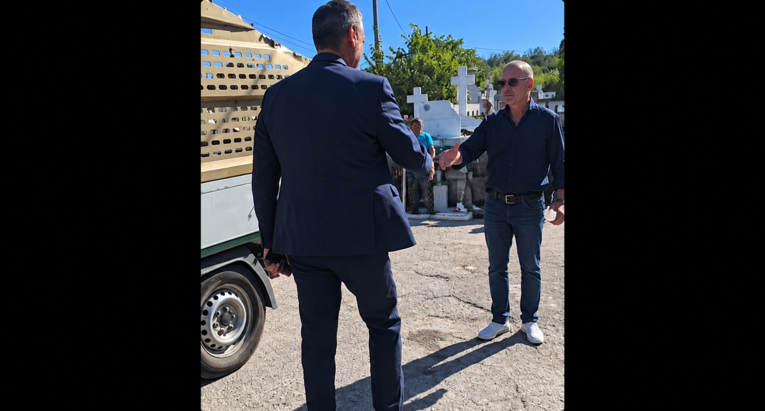 Two men shake hands beside a beige truck in a sunny cemetery with white crosses in the background, wearing smart and casual attire.