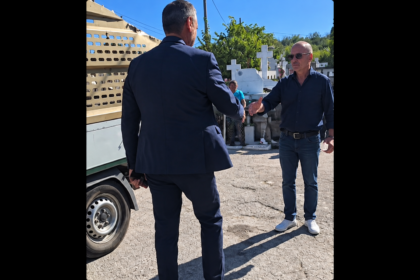 Two men shake hands beside a beige truck in a sunny cemetery with white crosses in the background, wearing smart and casual attire.