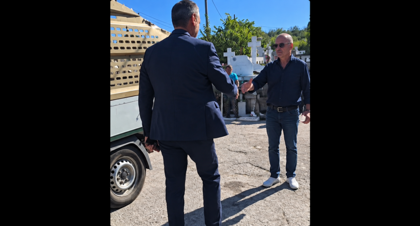 Two men shake hands beside a beige truck in a sunny cemetery with white crosses in the background, wearing smart and casual attire.