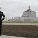 A man in formal navy dress uniform stands with hands on hips outside a fenced military facility with a US flag in view.