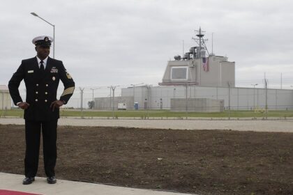 A man in formal navy dress uniform stands with hands on hips outside a fenced military facility with a US flag in view.
