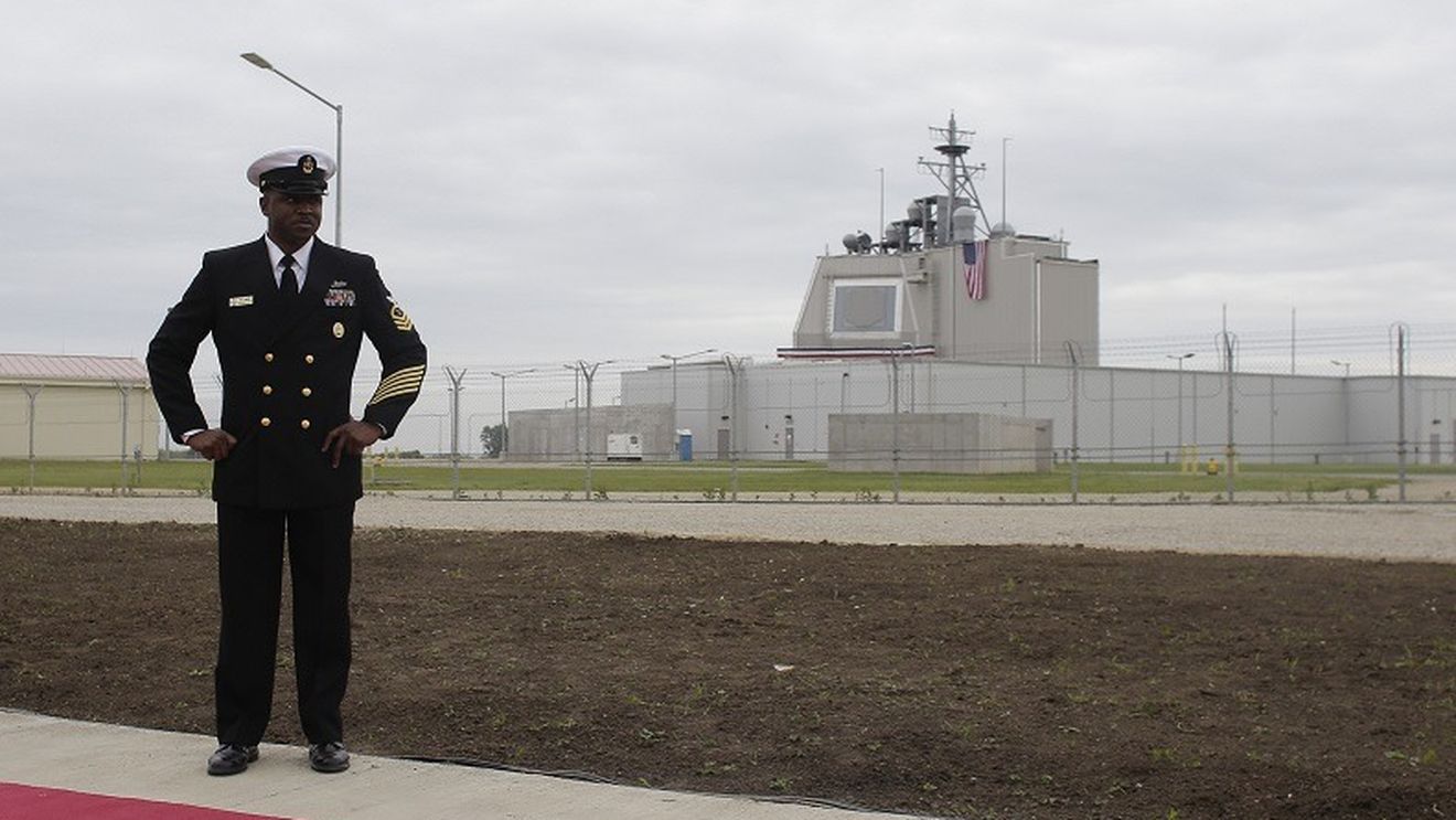 A man in formal navy dress uniform stands with hands on hips outside a fenced military facility with a US flag in view.