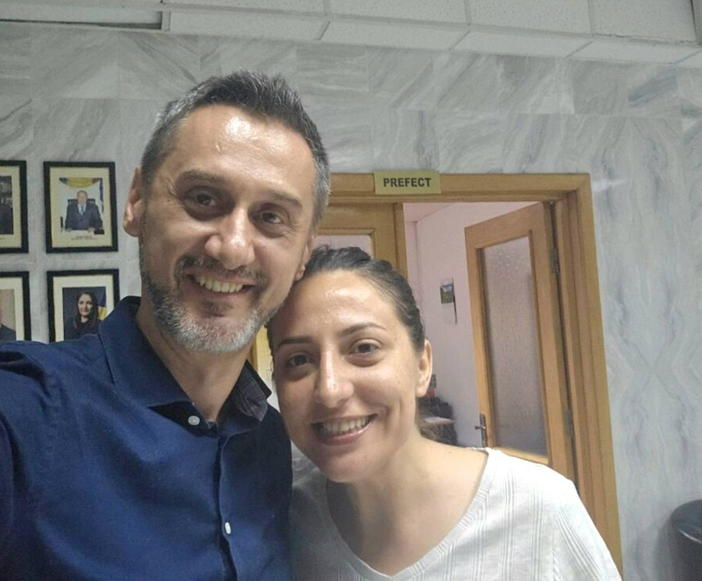 Two smiling adults posing for a selfie indoors, with a door labeled 'Prefect' in the background.