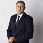 Professional man in a dark navy suit and blue tie, standing with hands clasped.