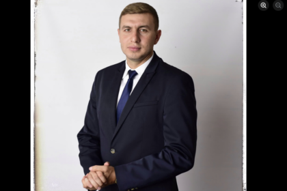 Professional man in a dark navy suit and blue tie, standing with hands clasped.