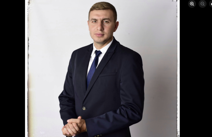 Professional man in a dark navy suit and blue tie, standing with hands clasped.