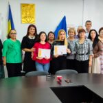 Twelve adults posing in a meeting room, two young women in front holding diplomas while others smile behind them, with flags and a gold emblem on the wall.