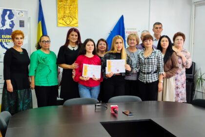 Twelve adults posing in a meeting room, two young women in front holding diplomas while others smile behind them, with flags and a gold emblem on the wall.