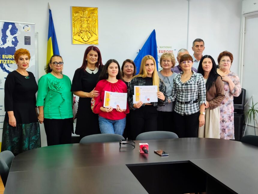 Twelve adults posing in a meeting room, two young women in front holding diplomas while others smile behind them, with flags and a gold emblem on the wall.