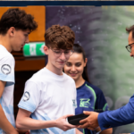Man in a blue suit presents an award to a young man in a white t-shirt at a ceremony, with others watching nearby.