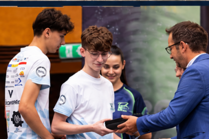 Man in a blue suit presents an award to a young man in a white t-shirt at a ceremony, with others watching nearby.