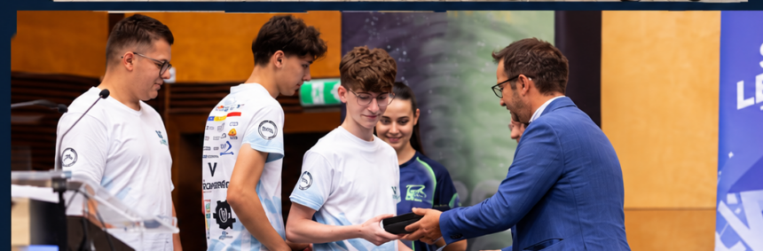 Man in a blue suit presents an award to a young man in a white t-shirt at a ceremony, with others watching nearby.