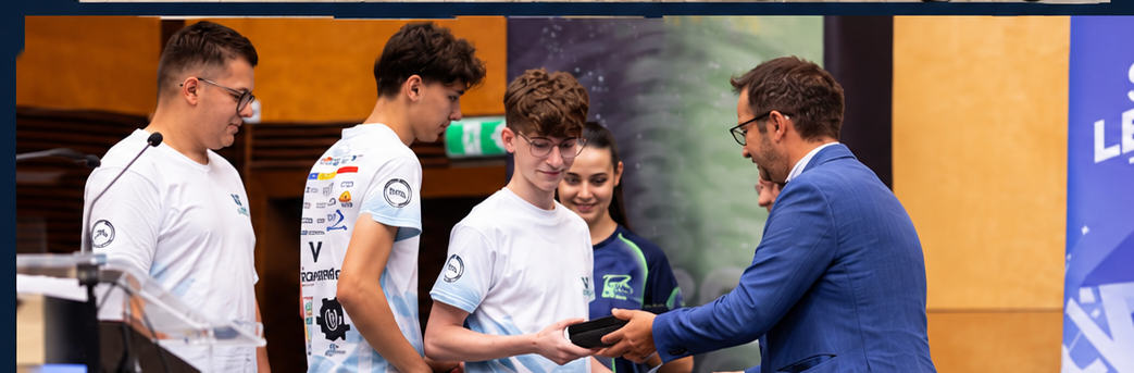Man in a blue suit presents an award to a young man in a white t-shirt at a ceremony, with others watching nearby.