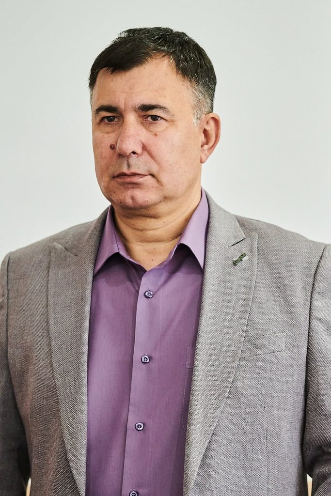 Portrait of a man in a gray blazer and purple shirt against a light background, looking slightly to the side with a neutral expression.